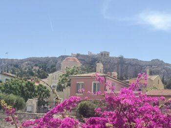 Rosa Bougainvillea im Vordergrund, pastellfarbene Gebäude in der Mitte und die Akropolis von Athen auf einem felsigen Hügel unter einem klaren blauen Himmel während der Heldinreise 2025 in der Plaka